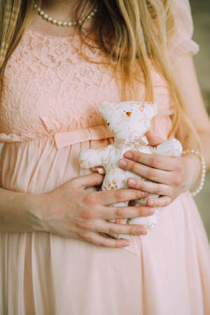 A pregnant woman in a delicate pink dress and a pearl necklace holds a white teddy bear in her handsの写真素材