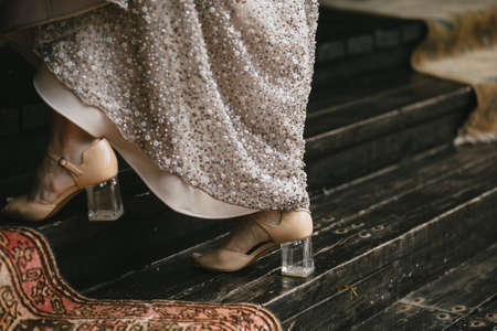 Beige shoes with a transparent heel on the brides legs go along the wooden staircase, evening dress with embroidery. Horizontalの写真素材