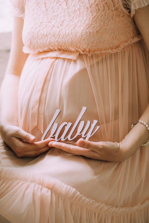 A pregnant woman in a tender pink dress holding the wooden word baby with her hands. Belly close-upの写真素材