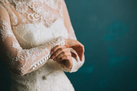 Female hands of the bride fasten buttons on the sleeve on a beautiful lace white wedding vintage dress close-up, morning preparation.の写真素材