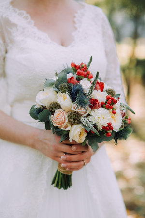 Beautiful elegant autumn wedding bouquet of white roses and red flowers in the hands of the bride in white wedding dress. Verticalの写真素材