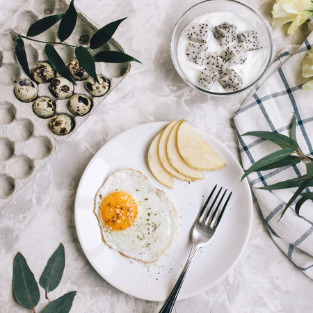 Breakfast of fried egg and pear on a white plate with a fork. In a glass bowl, yogurt and a dragon fruit, next to a tray of quail eggs, tablecloth and flowers on a concrete backgroundの写真素材