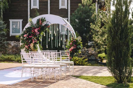 Wedding arch in the form of a white moon with floral compositions in a tropical style with leaves of a monster decorated with light bulbs. Transparent chairs for a ceremony in the street in the summerの写真素材