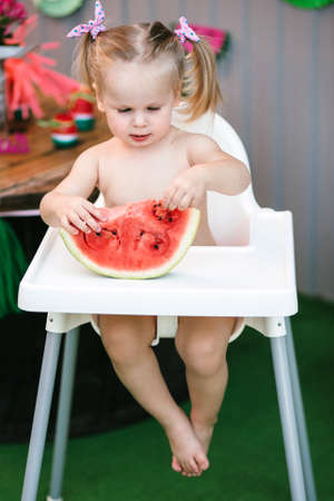A cute little girl baby blonde touches her hands a juicy piece of watermelon sitting in a children's chair with bare feet. Verticalの写真素材