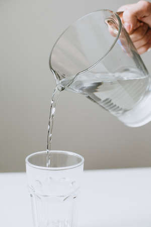 The hand holds a glass jug of water and pours into a transparent glass. Verticalの写真素材