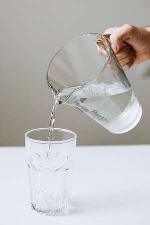 The hand holds a glass jug of water and pours into a transparent glass. Verticalの写真素材
