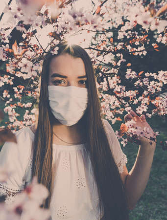 A young girl wearing a white mask for face protection against coronavirus. Standing in front of a blooming cherry blossom tree. Looking into the camera.の写真素材