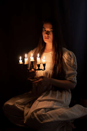 A young girl in an old white dress sitting and holding a candleabra in her hands. Dark background. Scary horror concept.の写真素材
