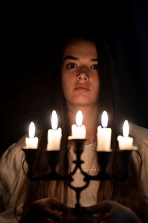 A young girl in an old white dress sitting and holding a candleabra in her hands. Dark background. Scary horror concept.の写真素材