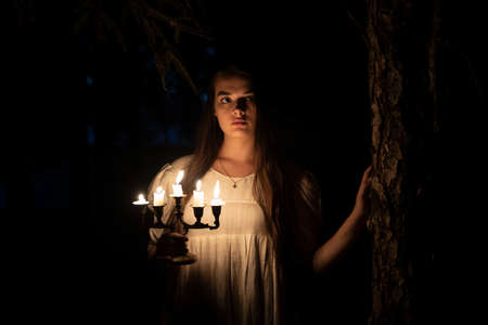 A young girl in an old white dress sitting and holding a candleabra in her hands. Dark background. Scary horror concept.の写真素材
