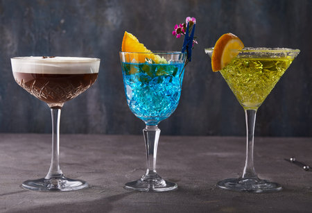 Three cocktails Espresso martini, Blue curacao and Thea standing in a row on a gray background.