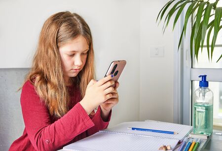 Distance learning online education. Schoolgirl studying at home with a laptop and doing school homework. Watching a task in a mobile phone. Concept Covid-19 or Coronavirusの写真素材