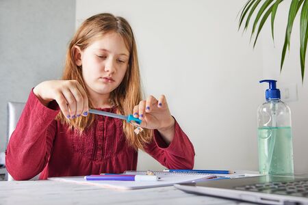 Distance learning online education. Schoolgirl studying at home with a laptop and doing school homework. Sharpen a pencil with a sharpener, selective focus. Concept Covid-19 or Coronavirusの写真素材