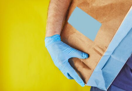 Delivery man employer in blue gloves hold craft paper bag with food, isolated on yellow studio background.の写真素材