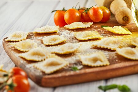 Tasty raw ravioli with flour, cherry tomatoes, sunflower oil and basil on a light wooden background. The process of making Italian ravioli. Copy space. Side view.の写真素材