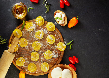 Tasty raw ravioli with flour, cherry tomatoes, sunflower oil and basil on a dark background. The...