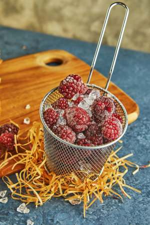 Frozen blackberries with ice in a metal mesh dish on a wooden board and with hay on a blue...