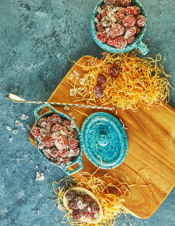 Frozen blackberries with ice in a blue glassware on a wooden board and with hay on a blue...