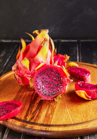 Dragon Fruit - Pitahaya Cut and one whole on a wooden stand against a black wooden background.
