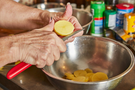 Cooking at home in the kitchen according to recipe from the Internet. A woman cuts potato with knife into slices.の写真素材