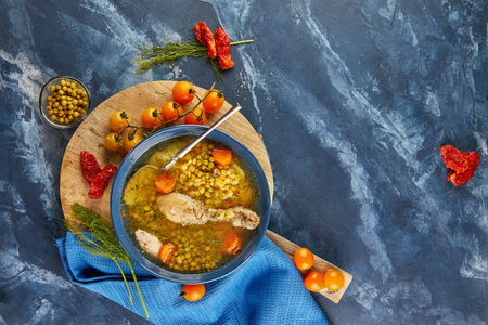 Pea soup with chicken, carrots, tomatoes and herbs in a deep plate on wooden board with napkin on blue backgroundの写真素材