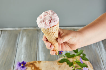Hand holding waffle cone with ice cream on gray background with flowers