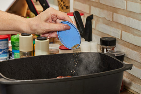 Chef adds ground black pepper to piece of beef fried in rectangular pan on gas stove. French gourmet...