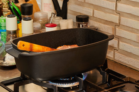 Piece of meat is fried with onions in wine sauce in rectangular pan over fire on gas stove. wooden...
