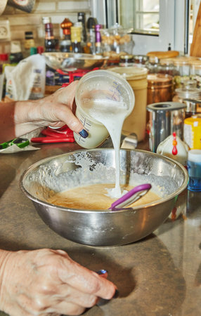 Chef in the home kitchen pours milk from measuring glass into pot with ingredients.