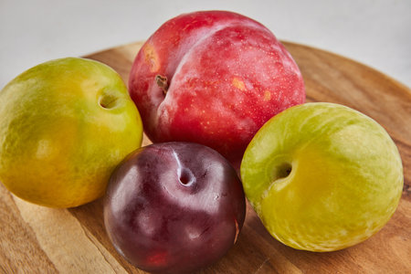 A variety of fresh plums arranged on a rustic wooden stand against a clean and elegant white marble background. The colorful fruits offer a bountiful display of natural, healthy goodness.の写真素材