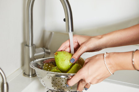 A woman is washing a handful of pears under running water in a kitchen sink. The pears are green and...