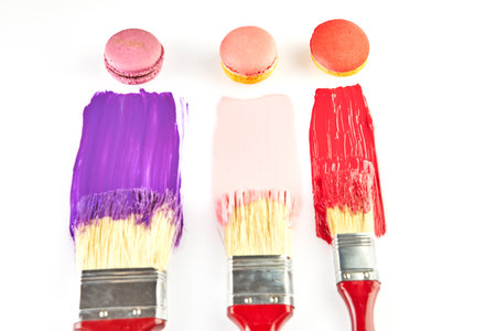 Three paintbrushes with red handles, each dipped in colorful paint, with matching macarons on a white background.の写真素材