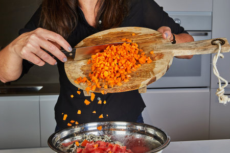 Close-up of hands using a knife to throw fresh carrots from a wooden cutting board into a pan in the...
