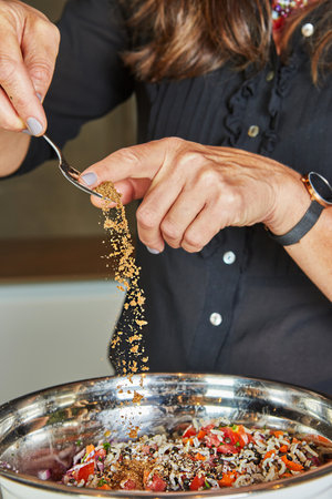 Close-up of a person sprinkling ground spices over a colorful salad in a stainless steel bowl in the kitchen.の写真素材