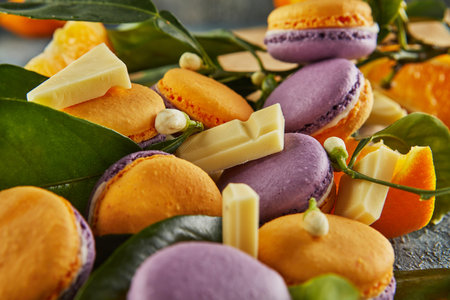 Vibrant yellow and purple macarons displayed with orange slices, green leaves, and white chocolate chunks on a rustic wooden board.の写真素材