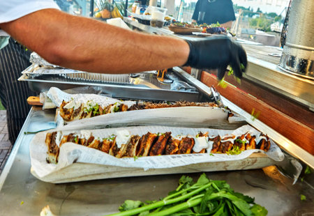 A chef is preparing a long sandwich with various ingredients at an outdoor event, surrounded by...