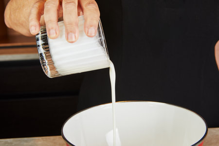 A hand pouring milk into a white mixing bowl, preparing ingredients for a recipe in a modern kitchen setting.の写真素材