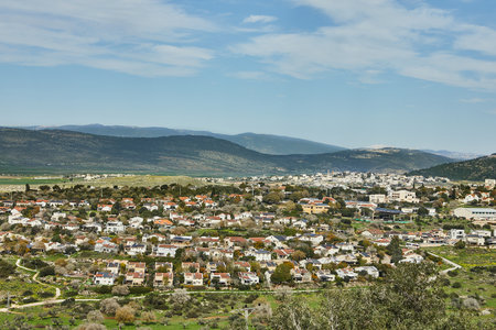 A panoramic view of a rural town nestled among rolling green hills, under a bright blue sky, showcasing nature and human settlementの写真素材