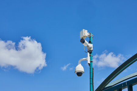 Urban security cameras and monitoring devices installed on a green pole above a public structure, set against a bright blue sky.の写真素材