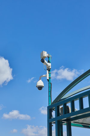 Urban security cameras and monitoring devices installed on a green pole above a public structure, set against a bright blue sky.の写真素材