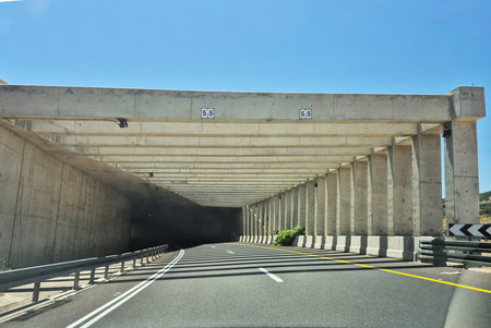 A modern concrete tunnel entrance with a 5.5m clearance height, casting strong shadows across the sunny mountain roadの写真素材