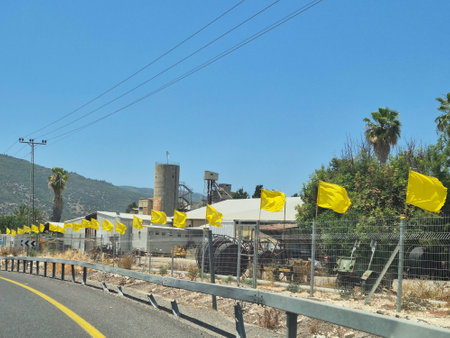 Yellow flags line a roadside fence beside an industrial building, with palm trees and forested hills under clear blue skiesの写真素材