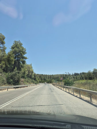 A straight country road leads through gentle hills and forested areas beneath a wide blue sky, viewed from a moving vehicleの写真素材