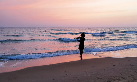 A person plays violin alone by the sea during a vibrant sunset, surrounded by waves and golden reflectionsの写真素材