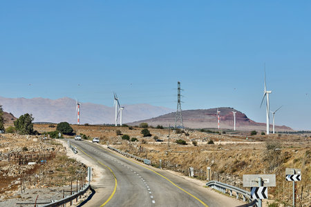 Winding road through a rural landscape with wind turbines and power lines beneath a clear blue sky.