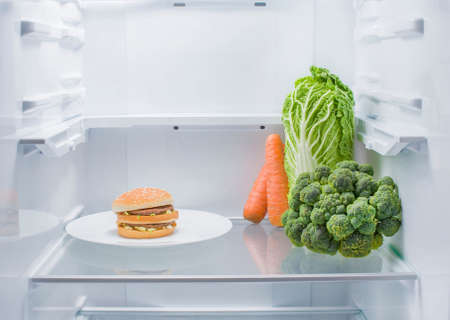 A hamburger on a plate and fresh vegetables opposite each other in an empty refrigerator. The choice between healthy foods and junk foodsの写真素材