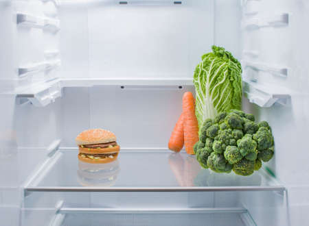 Hamburger and fresh vegetables opposite each other in an empty refrigerator. The choice between healthy foods and junk foodsの写真素材
