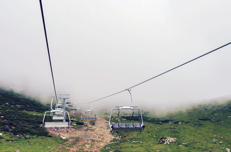 A lot of empty chairs of an open cableway in the mountains against a background of dense fog and...