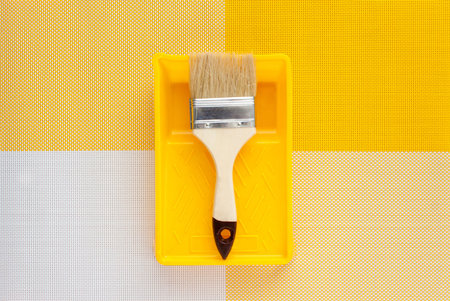 Paint brush and yellow paint tray on a symmetrical light yellow and white background. In the center