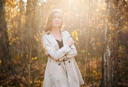 Beautiful, girl in a raincoat smiles in the autumn forest at sunset. Flare. Yellow foliageの写真素材